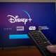 A television showing the Disney+ logo with Disney Pixar Marvel Star wars and National Geographic also labeled. A person points a remote toward the screen.